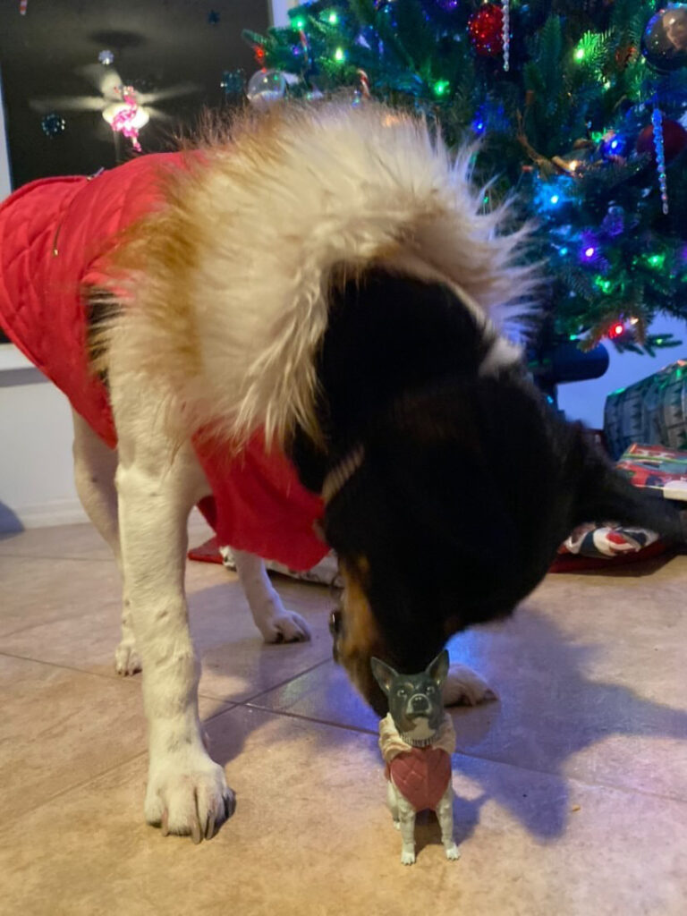 A dog figurine dressed in a holiday sweater placed under a decorated Christmas tree.